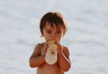 Repas de bébé en voyage : mes astuces pour des vacances sans stress Repas de bébé en voyage : un bébé boit son biberon à la plage pendant les vacances d'été
