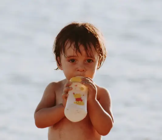 Repas de bébé en voyage : mes astuces pour des vacances sans stress Repas de bébé en voyage : un bébé boit son biberon à la plage pendant les vacances d'été